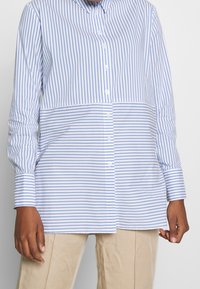 Randig skjorta med knappar i ljusblått och vitt, med långa ärmar, krage och rak passform. Materialet verkar mjukt och lätt.