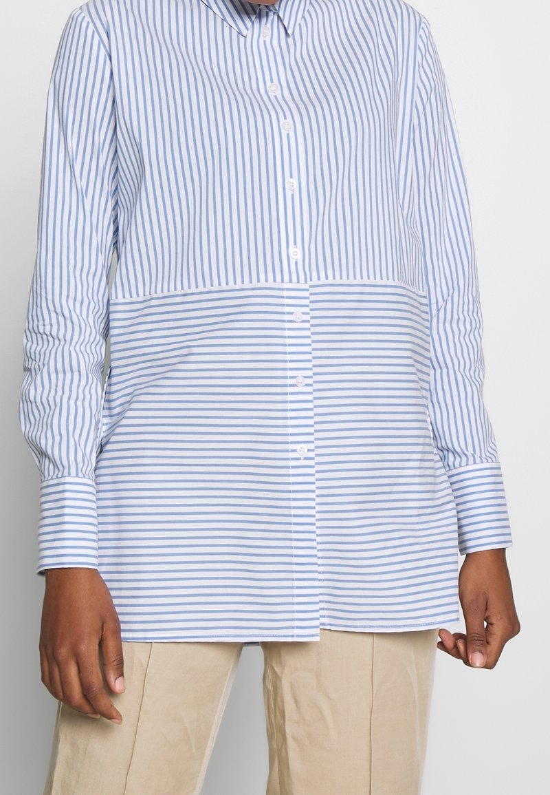 Randig skjorta med knappar i ljusblått och vitt, med långa ärmar, krage och rak passform. Materialet verkar mjukt och lätt.