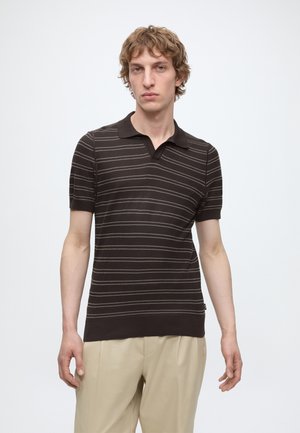 Man draagt een donkerbruine polo met korte mouwen met dunne horizontale strepen en beige broek, staand tegen een effen lichte achtergrond.