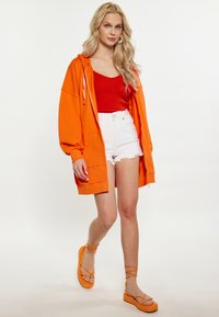 Felpa arancione brillante con zip sopra una canottiera rossa a coste, abbinata a pantaloni corti bianchi sfilacciati e sandali arancioni con cinturini. Tessuti morbidi e vestibilità rilassata.