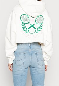 Witte cropped hoodie met grafische afbeelding van groene tennisrackets en bladeren op de achterkant. Draagt bijpassende lichtblauwe, hoge taille jeans met twee achterzakken.