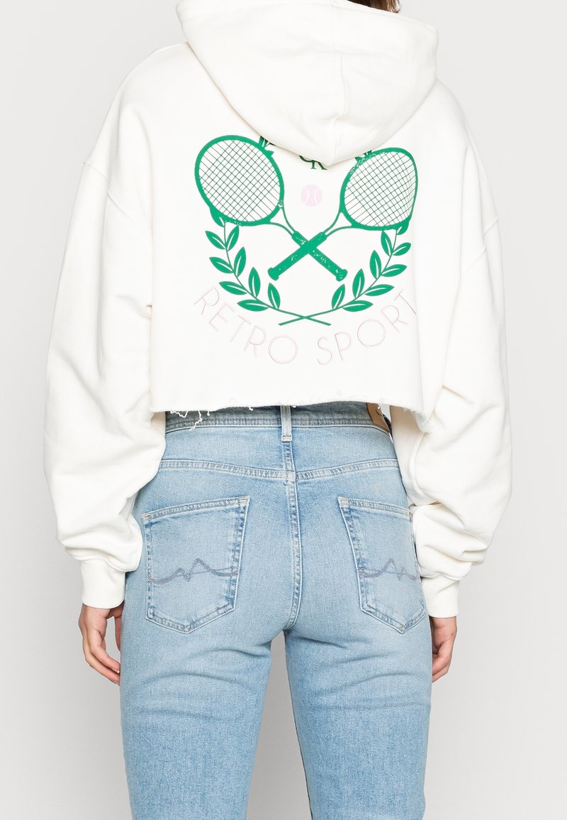 Witte cropped hoodie met grafische afbeelding van groene tennisrackets en bladeren op de achterkant. Draagt bijpassende lichtblauwe, hoge taille jeans met twee achterzakken.