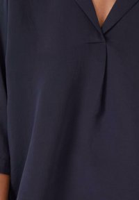 Blouse bleu marine avec un décolleté en V, des manches courtes et un tissu doux et texturé avec un léger éclat. Comprend un détail de pli central.
