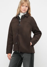 Bruine jas met een zachte afwerking, een kraag, knopen aan de voorkant, twee zijzakken en een losse pasvorm. Draagt over een geruit overhemd en grijze broek.