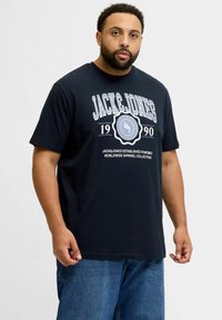 Tumedat sinine puuvillane t-särk, mis sisaldab julget valget teksti "JACK & JONES 1990" ja ümmargust logo graafikat, mis on selle all keskel. Mugav lõige.