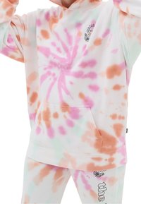 Personne portant un sweat à capuche blanc et un pantalon avec des motifs tie-dye rose, orange et vert clair, ornés de petits graphismes de soleil et de papillon.