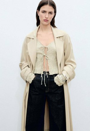 Cămașă bej cu trench peste un top scurt de culoare deschisă, texturat, cu legături. Pantaloni largi din denim închis. Accentele includ manșete cu volane și brățară.