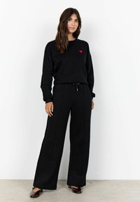 Fekete pulóver piros szív díszítéssel, fekete bő szárú nadrággal párosítva. Az outfit laza stílusú és puha textúrájú. A modellen sötét cipők vannak.