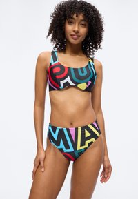 Γυναίκα που φοράει πολύχρωμο μαγιό δύο τεμαχίων με γεωμετρικό μοτίβο, στέκεται με ένα ελαφρύ χαμόγελο μπροστά σε ένα απλό ανοιχτό φόντο.