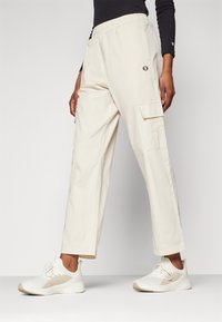 Bežové cargo kalhoty s elastickým pasem a bočními kapsami. Vyrobené z měkké tkaniny, s logem značky na levé straně. Dokončeno s bílými teniskami.