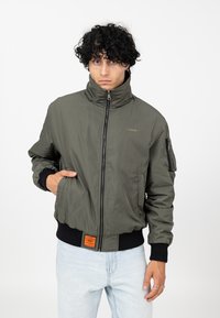 Veste bomber vert olive avec col montant, fermeture éclair, poches latérales et poignets côtelés. Elle comporte un petit logo et une étiquette orange.