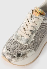 Sneaker con tomaia in finta pelle di serpente metallica, decorata con borchie argentate, lacci crema e un tallone riflettente. Suola chunky beige.