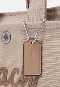 Sac cabas Coach en toile beige avec étiquette en cuir embossée "COACH NEW YORK" suspendue à une chaîne à billes argentée près du logo cousu et d'un fermoir métallique.