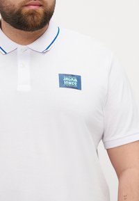 Camisa polo blanca con ribete azul en el cuello y un parche de logo rectangular azul en el pecho izquierdo que presenta el texto "JACK & JONES". Tejido texturizado.