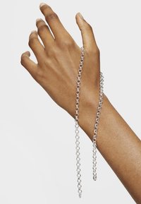 Pulsera de cadena plateada sobre una mano morena, con eslabones redondeados y un acabado pulido, con dos cadenas alargadas que se extienden hacia abajo.
