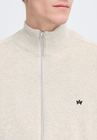 Suéter de color beige claro con cremallera y cuello alto, textura acanalada y un pequeño logotipo negro en el lado izquierdo.