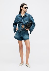 Sieviete valkā sasietu džinsa jaku un saskaņotas džinsa šortus, rokās tur melnu rokassomiņu, valkā melnus saulesbrilles un baltus slip-on apavus.