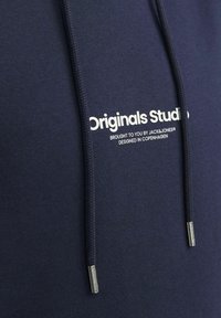 Námornícky modrá mikina s bielym textom "Originals Studio" na prednej strane, s viazacími šnúrkami s kovovými špičkami a mäkkou textúrovanou tkaninou.