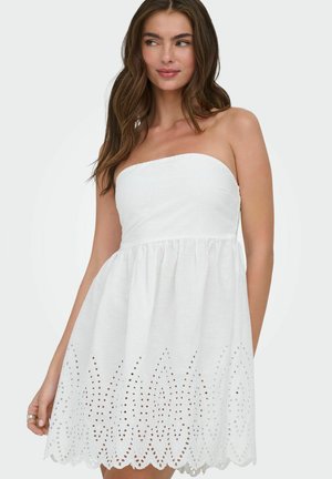 Kvinde med langt brunt hår iført en hvid stropløs kjole med øjestiks-skulderkant, ser let mod venstre mod en ensfarvet baggrund.