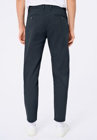 Chino gris foncé avec une coupe slim, deux poches arrière avec des boutons et des ourlets retroussés. Porte avec des chaussures blanches et des chaussettes blanches visibles.