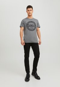 T-shirt in cotone grigio con testo grafico circolare nero, indossata con pantaloni neri e stivali neri con lacci, in piedi su uno sfondo neutro.