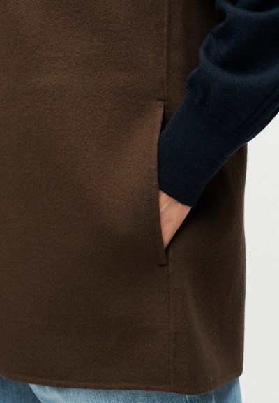 Καφέ ένδυμα με υφή υφάσματος και πλαϊνή τσέπη. Λεπτομέρεια με σκούρο μπλε πλεκτό μανίκι. Ορατή ραφή και ίσια ρεβέρ.