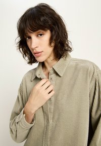 Person with short wavy hair wearing a beige corduroy button-up shirt, holding the collar with one hand against a plain light background.