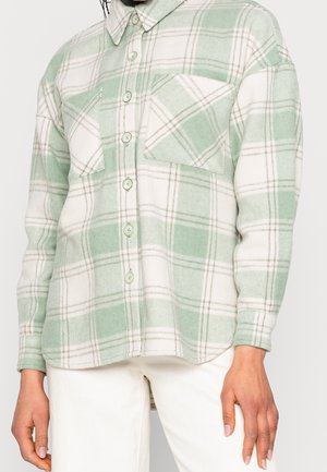 Vaaleanvihreä ja valkoinen ruudullinen nappipaita, jossa on kaksi rintataskua, pidettynä valkoisten housujen kanssa, kuvassa näkyy paita kaulasta lantioihin asti.