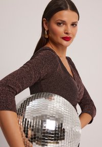 Brązowa błyszcząca sukienka z długimi rękawami, z dekoltem w kształcie litery V. Modelka trzyma dużą kulę dyskotekową. Proste złote kolczyki w kształcie kół dodają akcentu.