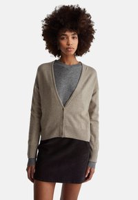 Kobieta o kręconych włosach, ubrana w beżowy kardigan z guzikami na szarym swetrze i czarnej spódnicy, stojąca na białym tle.