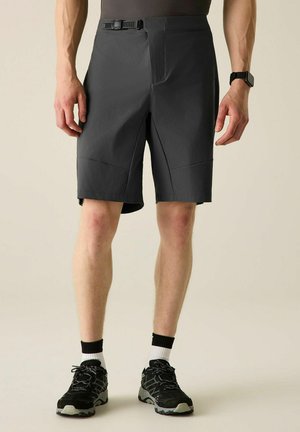 Mann trägt schwarze Sportshorts mit Gürtel, schwarz-weiße Socken und schwarze Laufschuhe und steht vor hellem Hintergrund.