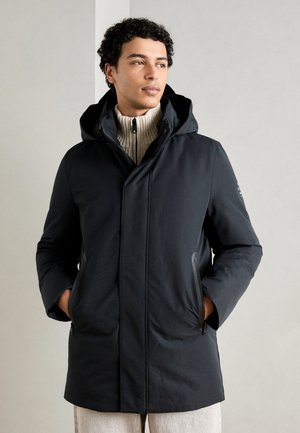 Chaqueta de plumas oscura con capucha, que cuenta con bolsillos delanteros con cremallera, un diseño elegante y una textura suave. Llevada sobre un suéter de canalé crema.