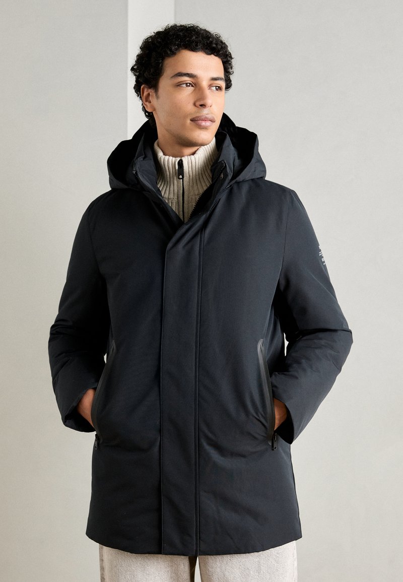 Chaqueta de plumas oscura con capucha, que cuenta con bolsillos delanteros con cremallera, un diseño elegante y una textura suave. Llevada sobre un suéter de canalé crema.