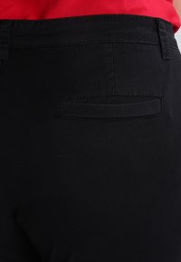 Un pantalon noir en tissu lisse et texturé. Il possède une poche arrière unique avec un rabat. Des coutures visibles le long des coutures.