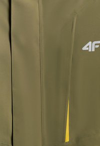Gros plan du tissu d'une veste vert olive avec des coutures verticales, une petite partie d'une doublure intérieure jaune et un logo blanc "4F".