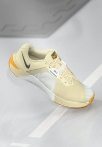 Ανοιχτό κίτρινο αθλητικό παπούτσι με πλέγμα, γκρι λεπτομέρειες, καουτσούκ σόλα και πορτοκαλί διακοσμήσεις. Διαθέτει ένα επιφανές λογότυπο στο πλάι.