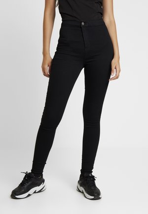 Woman wearing black high-waisted skinny jeans and black athletic sneakers, standing against a plain light background.