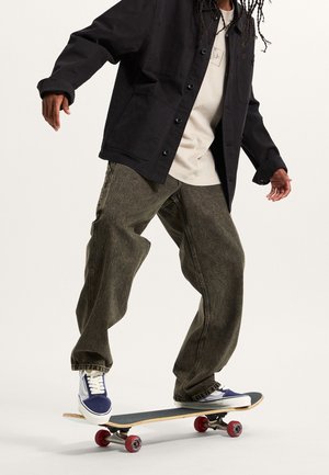Personne portant une veste noire, une chemise beige, un jean ample foncé et des baskets bleues, roulant en skateboard sur un fond blanc.