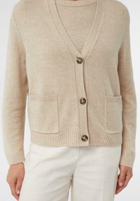 Beżowy kardigan z dekoltem w serek, z zapięciem na guziki, dwoma przednimi kieszeniami, ściągaczami przy rękawach i u dołu, wykonany z miękkiego materiału o fakturze.