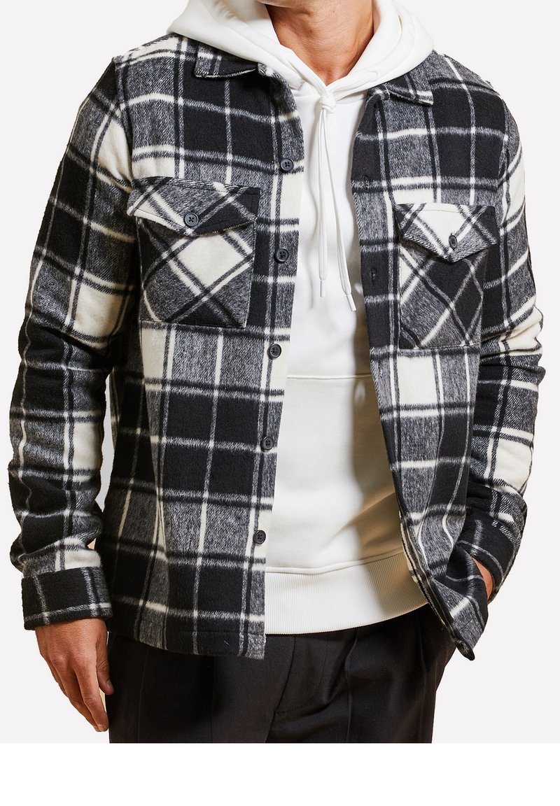 Chemise veste en flanelle à carreaux noir et blanc avec fermeture boutonnée, deux poches poitrine et une texture douce, portée par-dessus un sweat à capuche blanc.