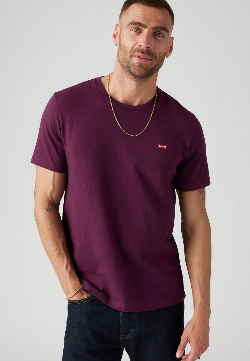 Burgundi T-shirt iš medvilnės, apvalios kaklo dizainas su trumpomis rankovėmis ir mažas raudonas Levi's logotipas krūtinėje, derinamas su tamsiomis džinsais.