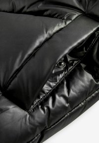 Veste doudoune noire avec un extérieur brillant et matelassé, des coutures visibles et une fermeture à boutons-pression. Le tissu semble doux et lisse.