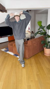 Grå fleecejakke med turkise detaljer, grå bukser med vide ben og sølvfargede joggesko. Fargerik teppe og teksturert oransje sofa i bakgrunnen.