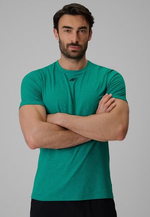 Mężczyzna o ciemnych włosach i brodzie stoi z założonymi ramionami, ubrany w zieloną sportową koszulkę z krótkim rękawem i czarne spodnie na szarym tle.