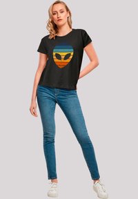 Black t-shirt featuring a colorful, retro alien graphic with horizontal stripes in blue, orange, and yellow. Lightweight, casual fit.