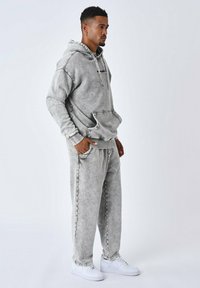 Sudadera gris con efecto de teñido anudado, con bolsillo frontal y cordón, combinada con pantalones de chándal a juego, completada con zapatillas blancas. Tejido suave y texturizado.