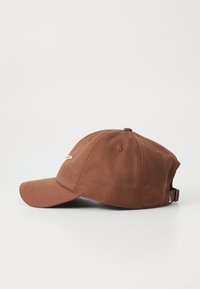 Brun seks-panel baseball cap med buet skygge, liten hvit brodert logo på fronten, og justerbar stropp bak på hvit bakgrunn.
