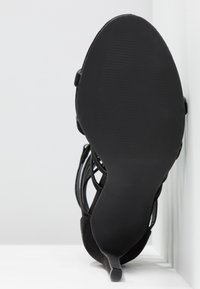 Chaussure à talon haut noire avec une tige en daim texturé, un design ouvert et un talon fin et pointu. La semelle est lisse avec des détails nervurés.