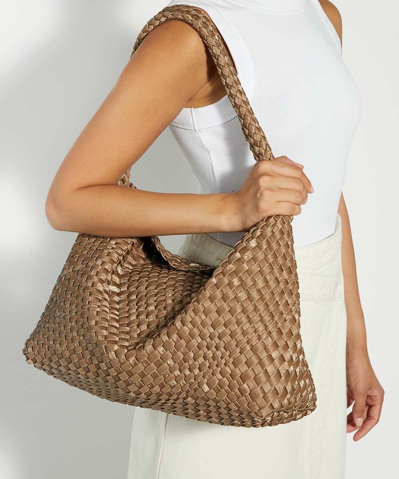 Femme tenant un grand sac à bandoulière tressé beige avec une épaisse bandoulière tressée, portant un haut blanc sans manches et une jupe de couleur claire.