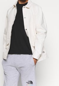 Light-colored, button-up jacket over a black shirt, paired with gray sweatpants featuring a logo. Cotton texture, casual design.